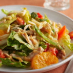 Chicken salad with peanut dressing on a white plate pictured on a wooden table with a fork and glass.