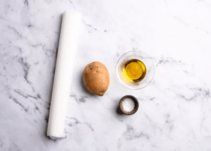 Parchment paper, olive oil in a cup, salt in a cup, and potato on a marble backdrop