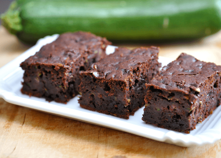 Three brownies on a white plate with a zucchini in the back